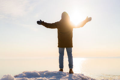 Rear view of man standing on snow by sea against sky during sunny day