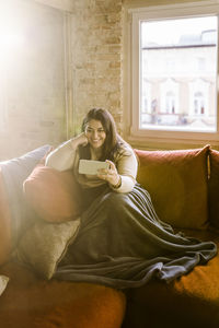 Smiling woman doing video call through smart phone while sitting on sofa at home