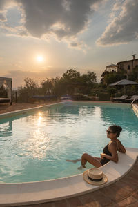 Woman sitting by swimming pool against sky during sunset