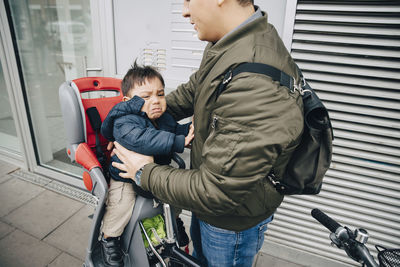 Midsection of father holding crying son sitting on bicycle seat at sidewalk in city