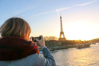 Rear view of woman photographing eiffel tower by seine river against sky during sunset