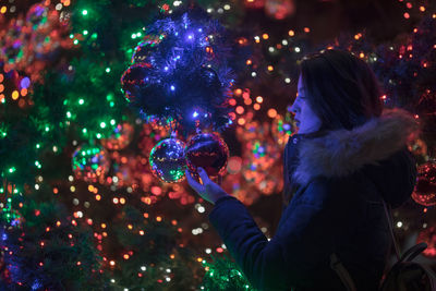 Woman holding illuminated christmas ornament on tree at night
