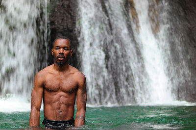 Portrait of shirtless man standing against waterfall