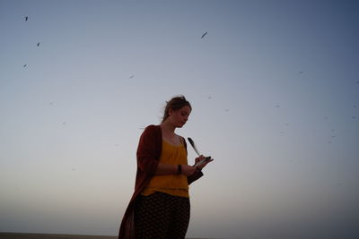 Full length of young woman using mobile phone against clear sky