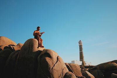 Low angle view of mid adult man playing guitar while sitting on rock formation against blue sky