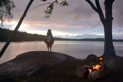 Rear view of woman standing at lakeshore against cloudy sky during sunset