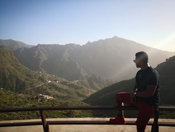 Man looking at mountains while sitting at observation point against clear sky