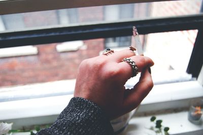 Cropped hand of woman smoking cigarette by window at home