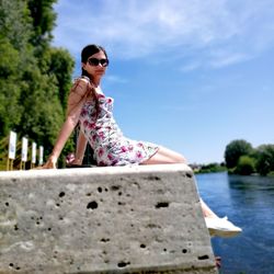 Side view of young woman relaxing on retaining wall by river during sunny day