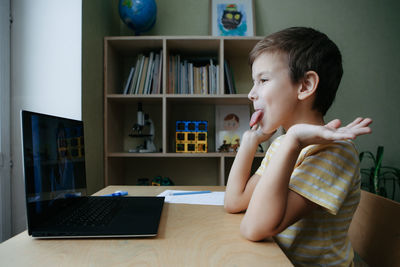 8 years old boy sit by desk with laptop and sticking out his tongue and fooling around. side view