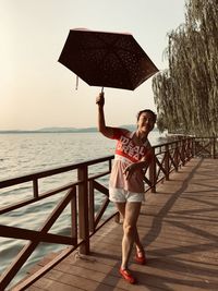 Full length of woman standing with umbrella by railing over sea against sky