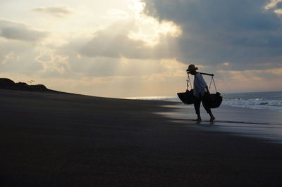 Vendor walking at beach against cloudy sky during sunset