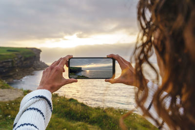 Cropped image of hand holding camera against sky during sunset