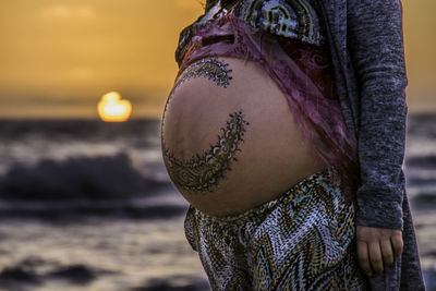 Midsection of pregnant woman with tattoo on stomach standing against sea during sunset