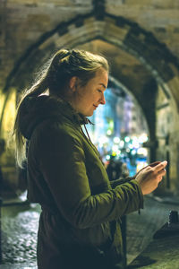 Side view of woman using phone while standing on street at night