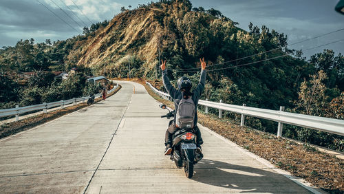 Rear view of man with arms raised sitting on motorcycle over mountain road