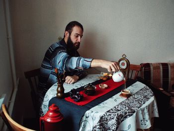 Man pouring tea while sitting at dinning table against wall