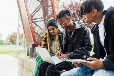 Smiling young man using laptop sitting by friends on wall