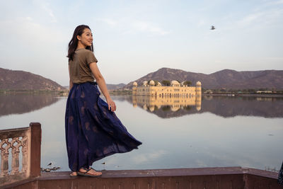Happy young asian woman looking away while standing on border near lake against jal mahal palace and mountains in jaipur, india