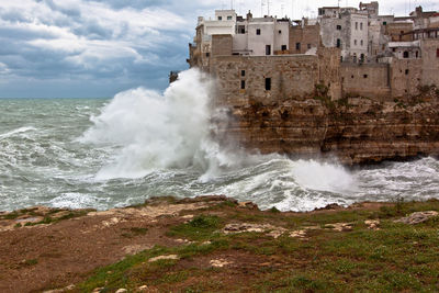 Sea waves splashing on buildings against cloudy sky