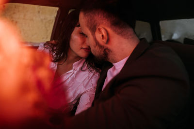 Newlywed couple kissing inside a car with red neon lights and bouquet flowers in foreground out of focus.