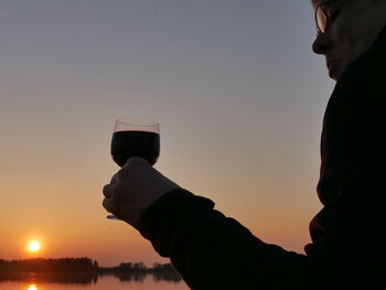 Midsection of man holding beer glass against sky during sunset