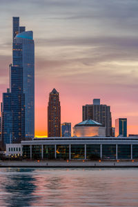 Modern buildings by lake against sky during sunset