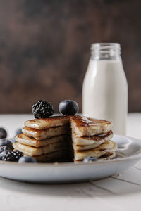 Still life of a stack of homemade pancakes with berries and syrup on a plate, and a bottle glass of milk in background.