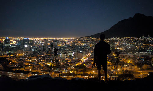 Rear view of silhouette man standing with tripods against illuminated cityscape