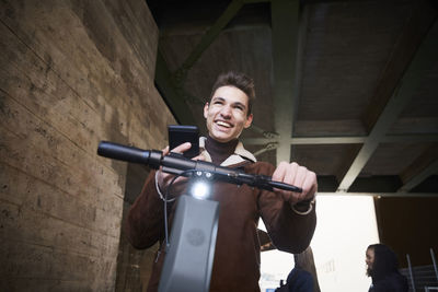 Happy teenage boy with electric push scooter looking away while holding mobile phone below bridge