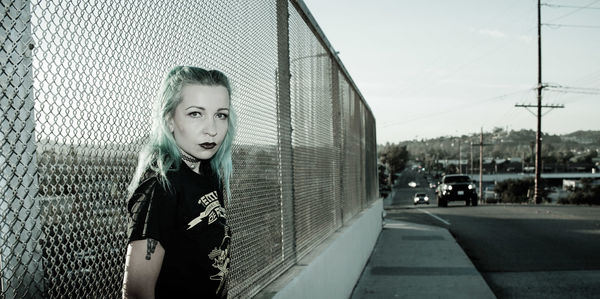 Portrait of female punk standing on sidewalk by road against fence