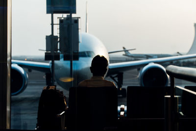 Rear view of man sitting at airport lobby against airplane