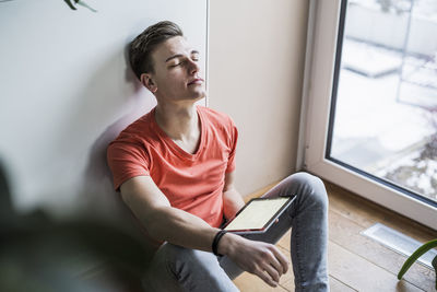 Young man with digital tablet relaxing while sitting against wall in living room