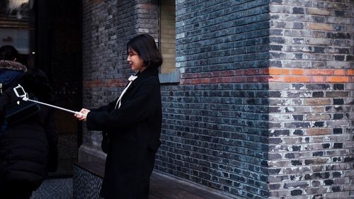 Side view of young woman taking selfie with smart phone while standing against brick wall