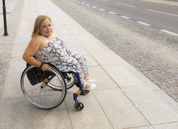 Smiling young pretty woman with short stature on wheelchair stands on bus stop outdoor waiting for