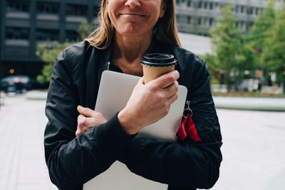 Midsection of mature businesswoman holding coffee cup with laptop in city