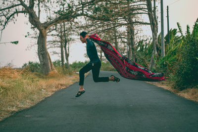 Side view of man holding scarf jumping on road in forest
