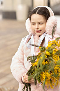 Portrait of a toddler girl in a warm pink coat and fur headphones with a bouquet of mimosa 