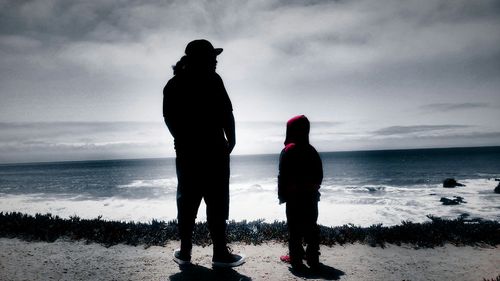 Rear view of father and son standing at beach against cloudy sky