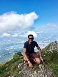 Portrait of young man wearing sunglasses while sitting on mountain against sky