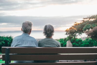 Rear view of woman sitting on bench against sky during sunset