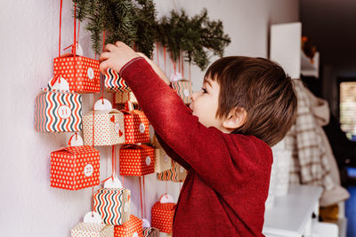 Little happy curious child opening first gift of handmade advent calendar hanging on wall. 