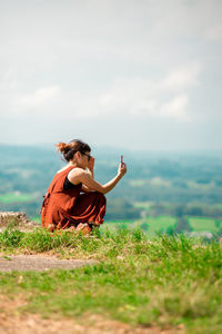 Side view of young woman using smart phone while sitting on mountain against sky