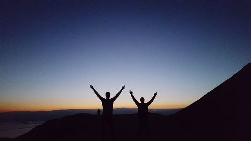 Silhouette friends with arms raised against clear blue sky