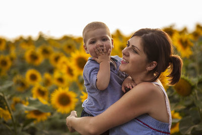Smiling mother carrying cute son while standing against sunflowers