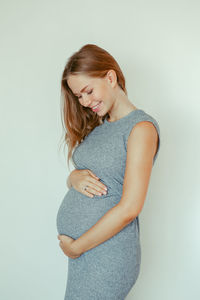 Pregnant young woman touching abdomen while standing against white background