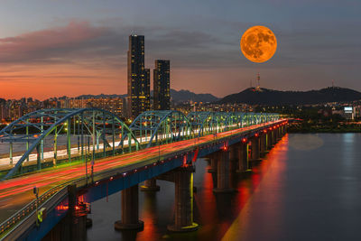 Illuminated bridge over river and buildings against sky during sunset