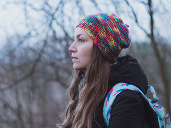 Young confident millennial woman long brown hair knitted hat black coat with backpack walks outside.