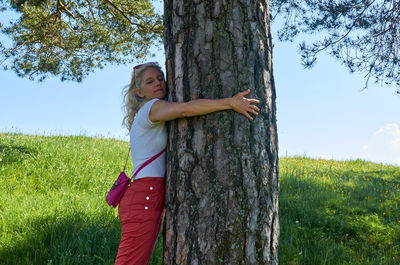 Woman standing by tree trunk on field against sky