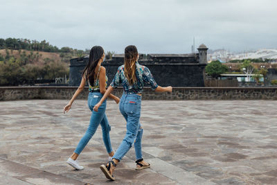 Side view of young slim girlfriends in trendy outfits and protective masks walking together on paved urban square during coronavirus outbreak